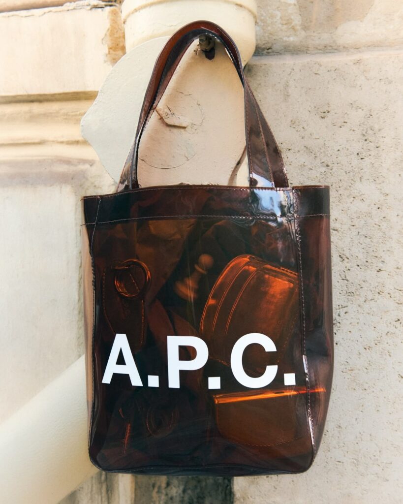 Sac transparent A.P.C. avec logotype blanc photographié devant une façade en pierre