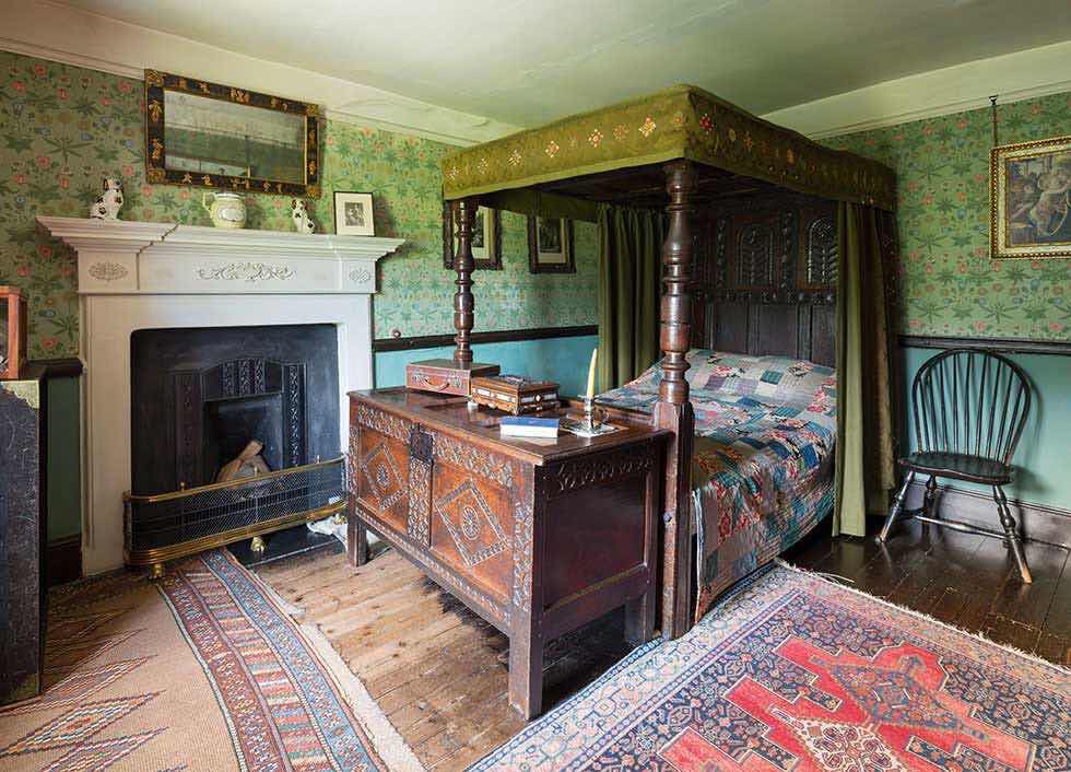 Chambre de Kelmscott Manor avec lit à baldaquin, cheminée et papier peint vert à motifs végétaux.