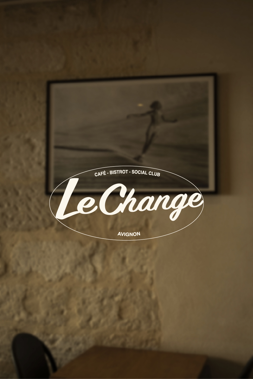 Logo blanc Le Change sur une photographie du mur en pierre et d’une table du restaurant.